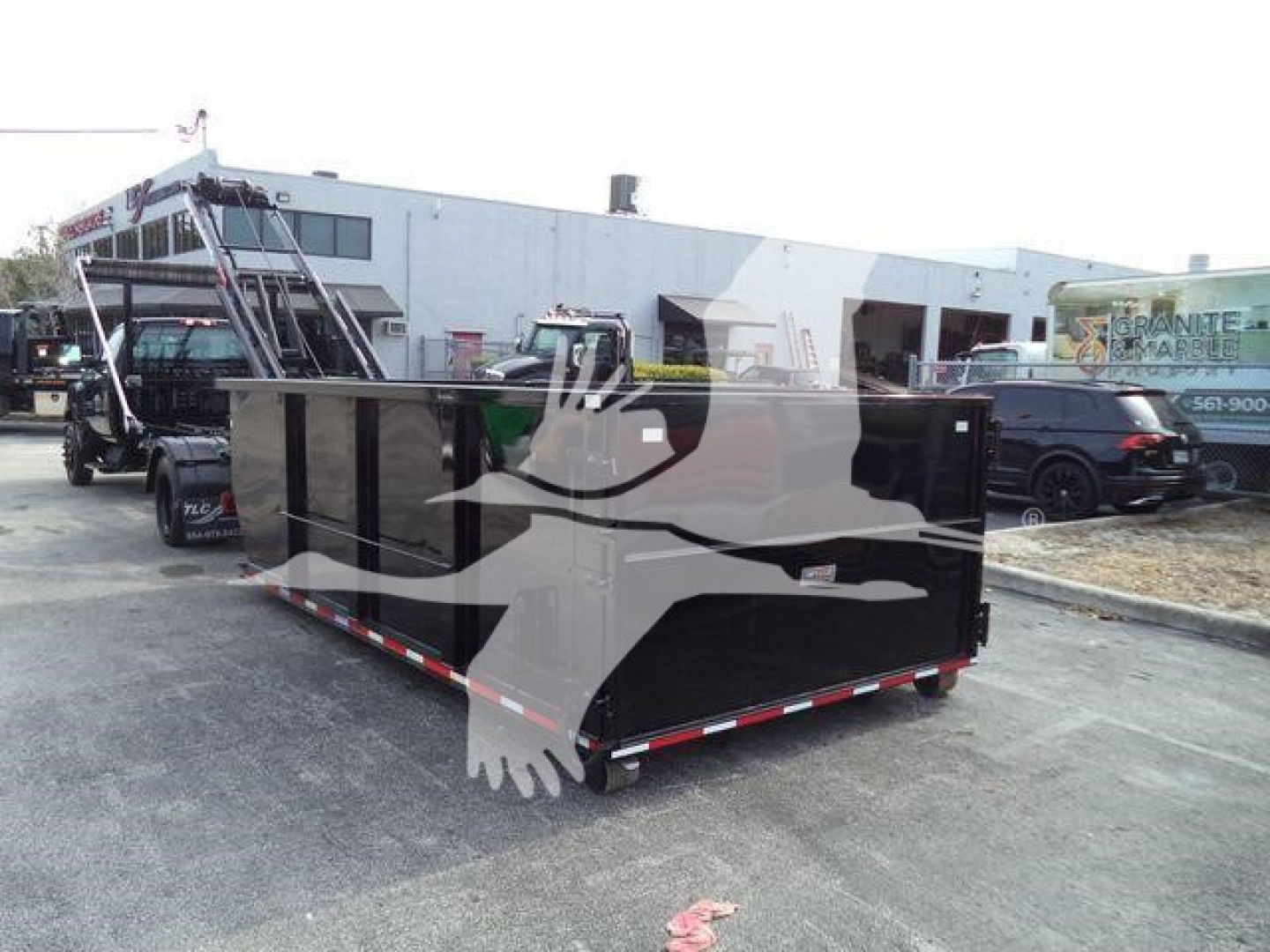 Rolloff truck bed trailer in a lot with a black long steel trailer, side rails, and white building in background.