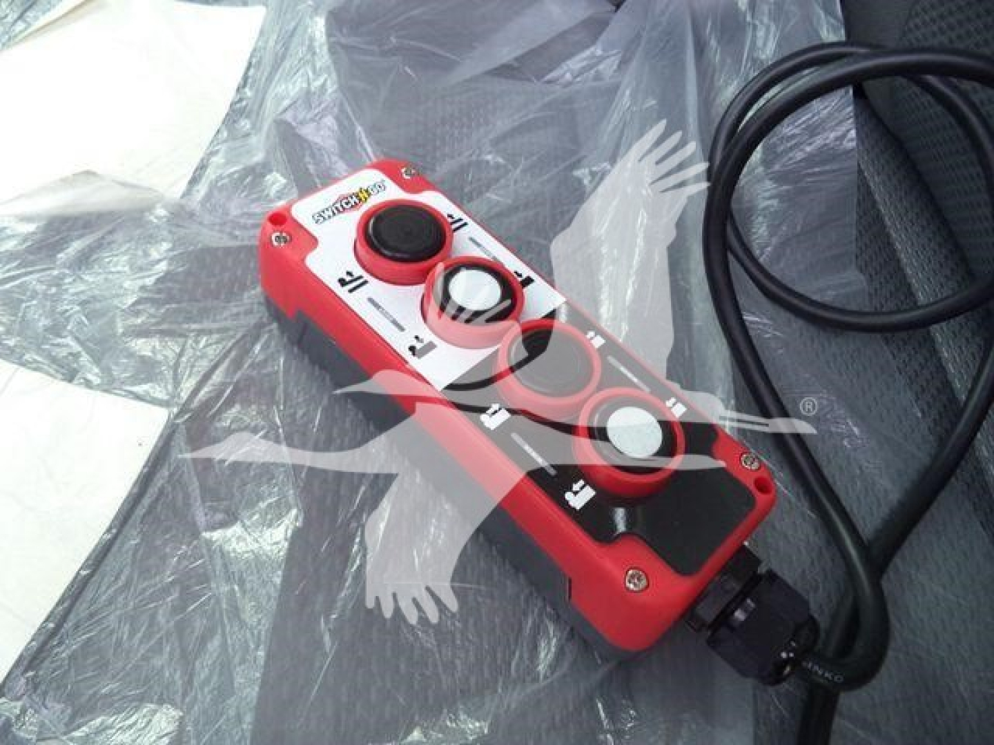 Remote control device with red housing and circular dials on a truck seat, showing control buttons and a black cable.