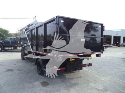 Truck flatbed trailer in a yard showing a black roll-off container mounted on a Silverado chassis with rear bumper and hydraulic hookups visible.
