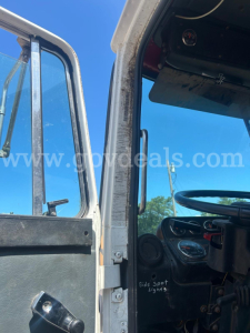 Cab interior of a truck showing steering wheel, dashboard, and open door frame from a left-hand side angle.