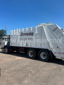 Front-left side of a garbage truck with a large white waste container. A banner on the side reads ‘Superior Waste Services’ with a phone number. The truck has multiple axles and black tires, under clear blue sky.