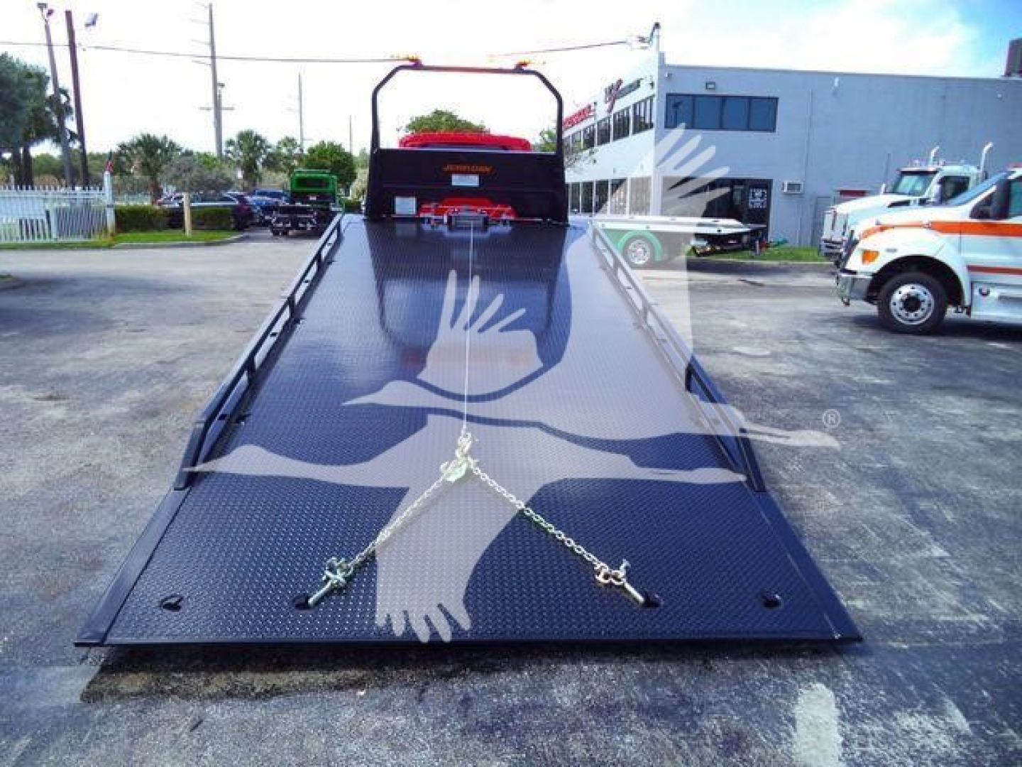 Flatbed tow truck deck with a diamond plate surface and a bird graphic, viewed from the rear, chains and hooks stretched across the bed, truck in a lot.