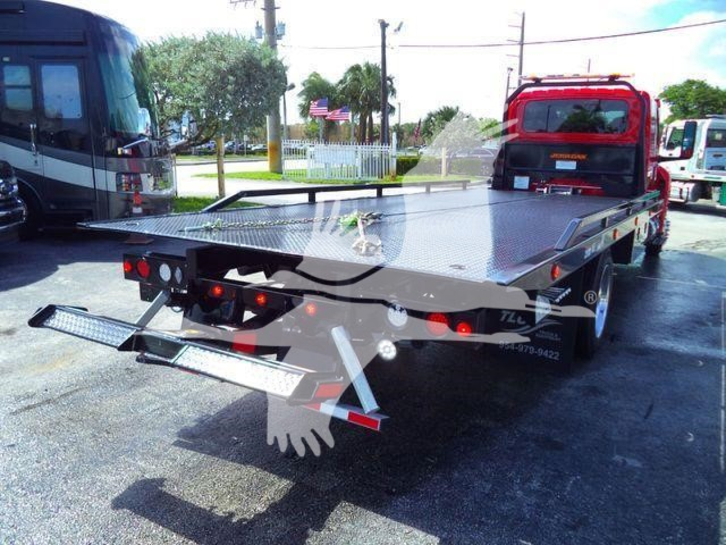 Flatbed tow truck car hauler with hydraulic rear ramps and reflective lights, viewed from rear left side.