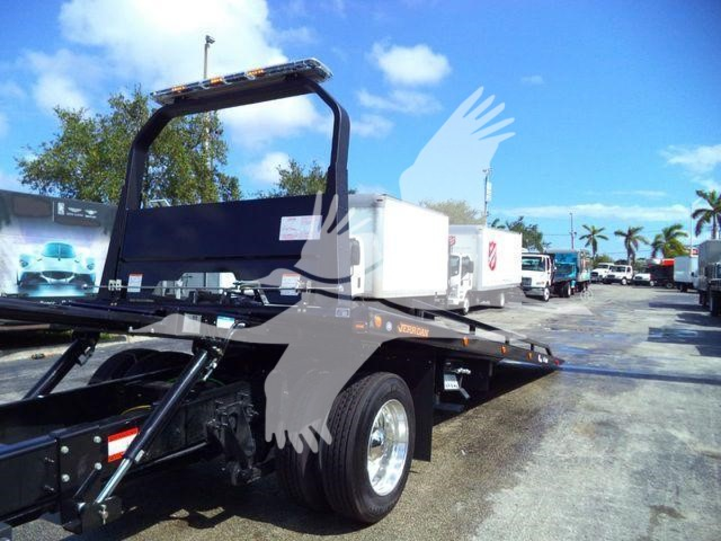 Tow truck rollback bed on a new International MV with dual rear wheels and a white storage box, dealership lot in background.