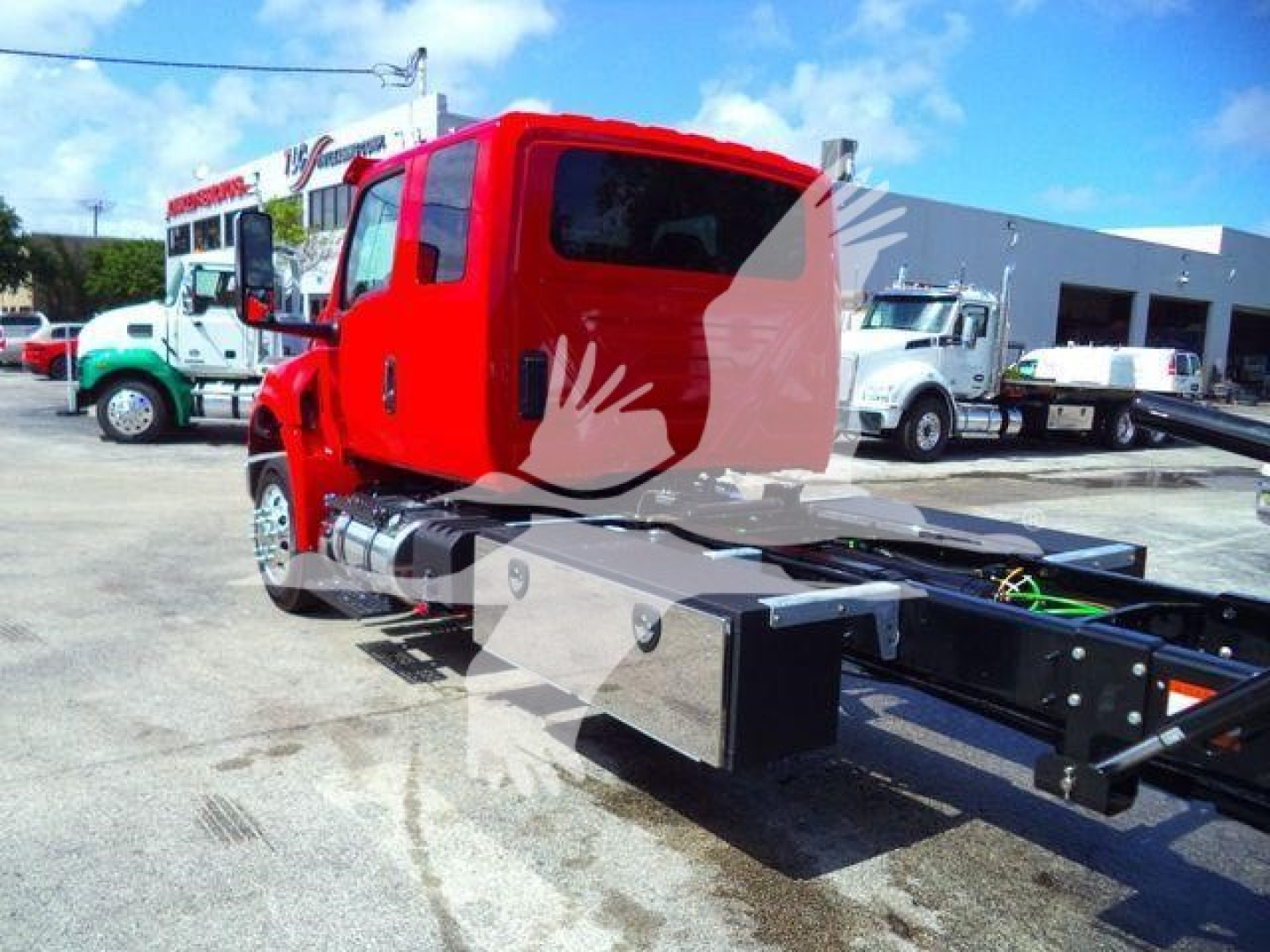 Truck chassis with red cab and flatbed undercarriage in a dealership lot; other trucks and a bus in background.