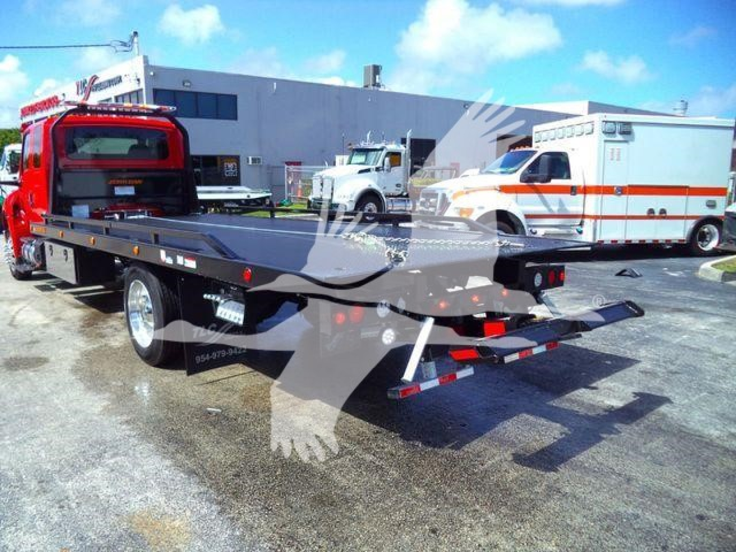 Truck rollback tow truck flatbed with red cab and extended wheel lift showing rear view and emergency vehicles in background.