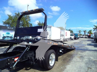 Tow truck rollback bed on a new International MV with dual rear wheels and a white storage box, dealership lot in background.