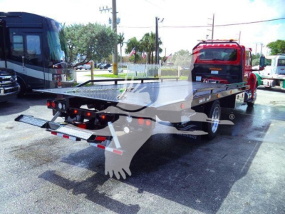 Flatbed tow truck with hydraulic ramps and drop-down loading deck, attached to a red semi-truck tractor in a dealership lot.