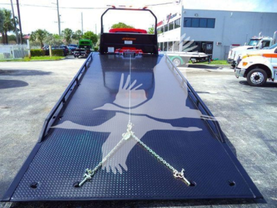 Flatbed tow truck ramp with diamond plate surface and a chain tie-down; background shows truck dealership lot with a white building and other heavy vehicles; text not clearly legible on the image.