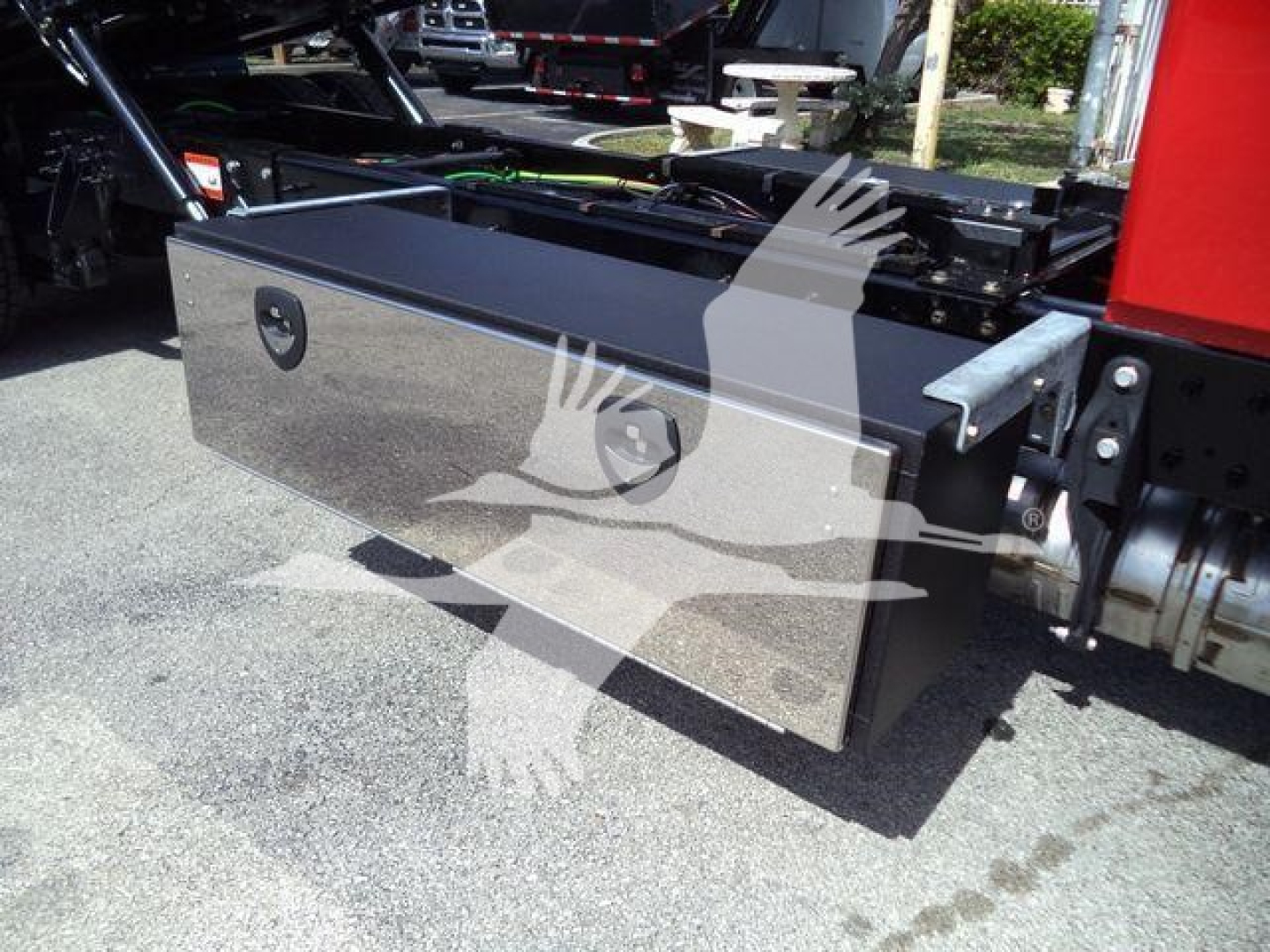 Vehicle equipment box mounted under truck with metallic diamond-plate dimensions, circular lock areas visible on front panel, hydraulic lines and chassis components in background.