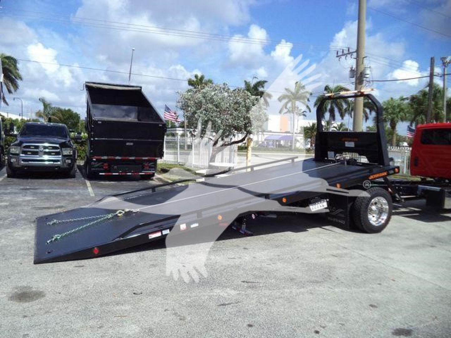 Flatbed rollback tow truck on a lot with other trucks in background, black flatbed with black cab and extended bed, blue skies.