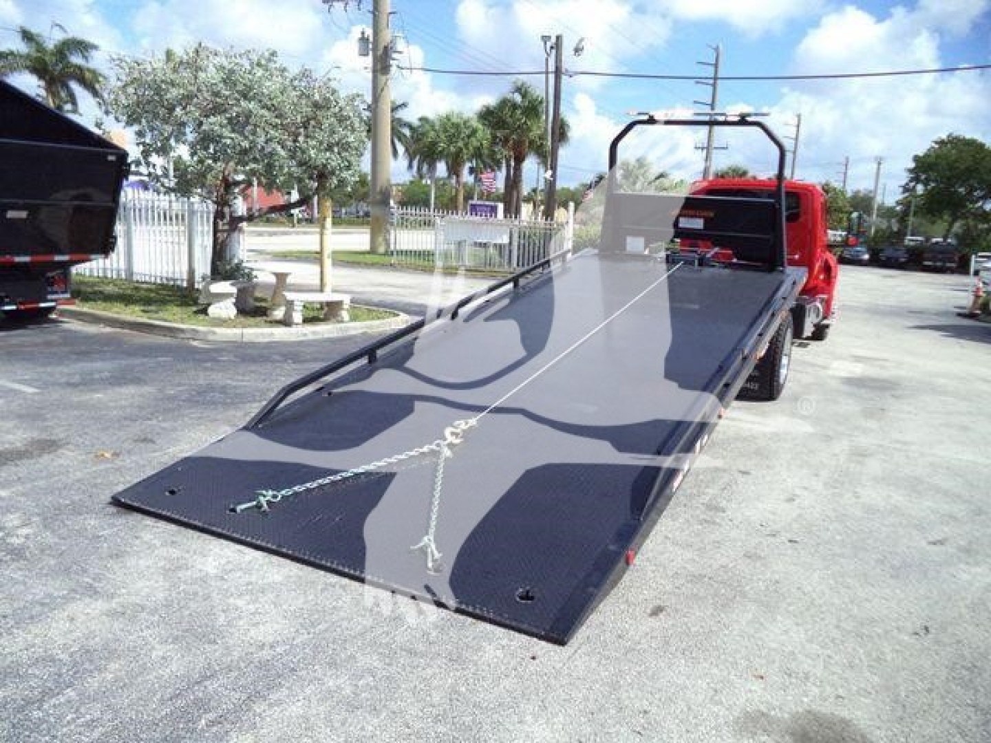 Truck bed with a black hydraulic rollback/shipping platform attached to a red truck, showing a lowered flat loading ramp and chain tie-downs.