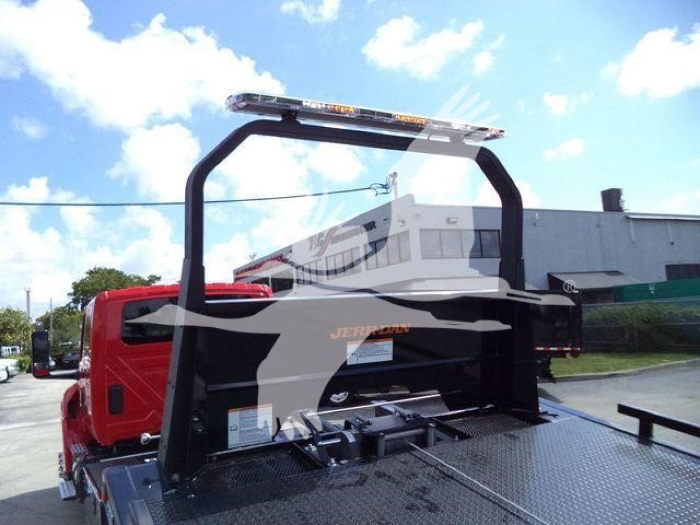 Truck rollback bed with metal diamond plate and a curved black rollover protective frame; red cab in background; warning light bar on top.