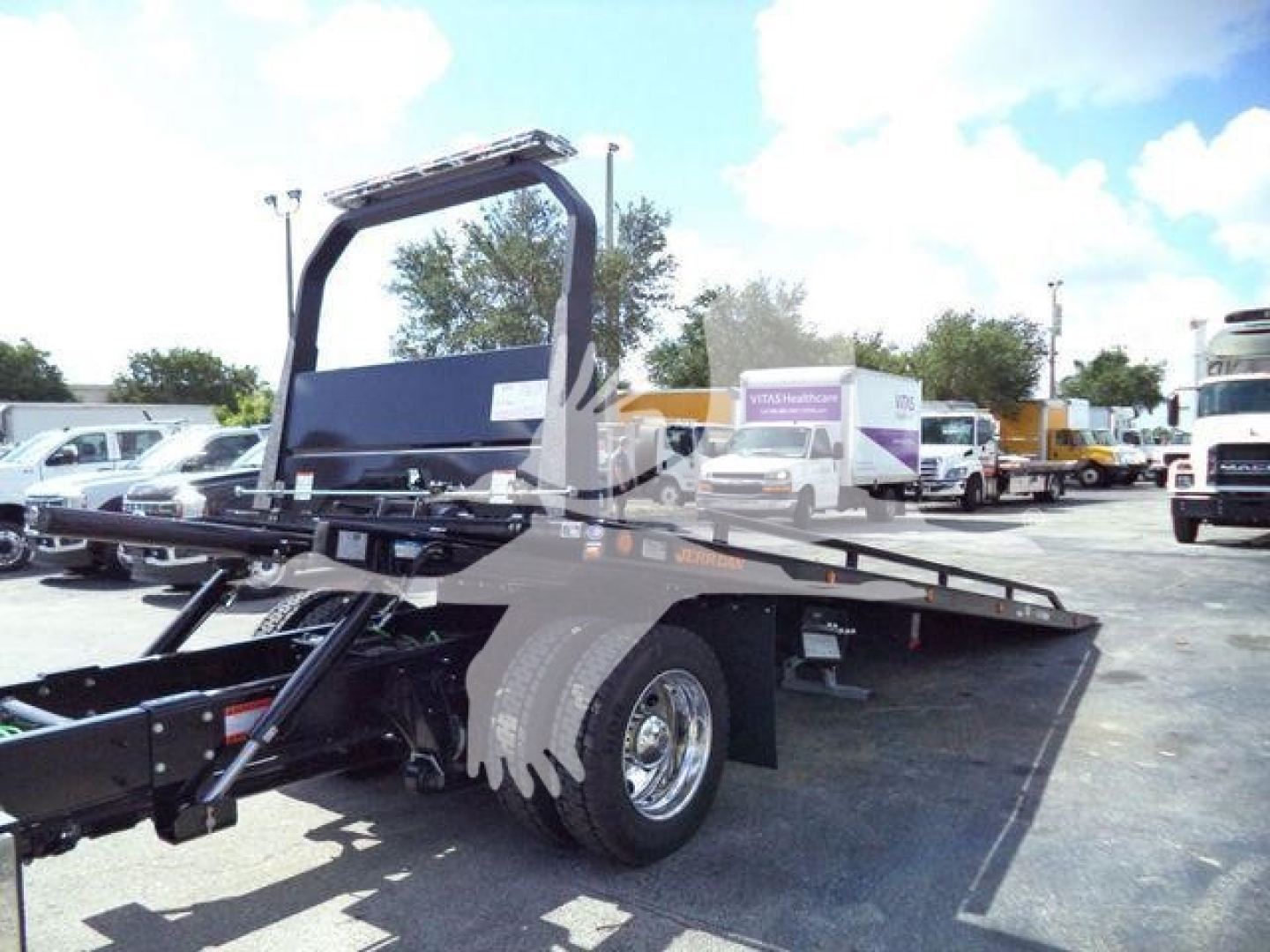Truck rollback bed with large steel frame and wheel, showing a flatbed and hoist mechanism; background includes other trucks and a clear sky.