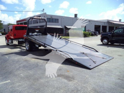 Flatbed rollback tow truck with a long metal ramp and black anti-slip surface. In the background is a white industrial building and a red truck to the left. The image contains reflections on the surface and a watermark across the center.