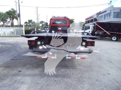 Rear view of a flatbed rollback truck with a red cab in a dealership lot. Visible are taillights, bumper, and reflective markers; other trucks are in the background.