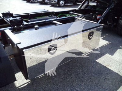 Toolbox/storage box mounted on a truck chassis, reflective metal surface with two circular black access ports, viewed in a truck yard.