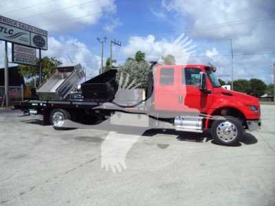 Tow truck with a red cab and flatbed rollback body, showing a black dumpster container on the bed. Clear sky with some clouds.
