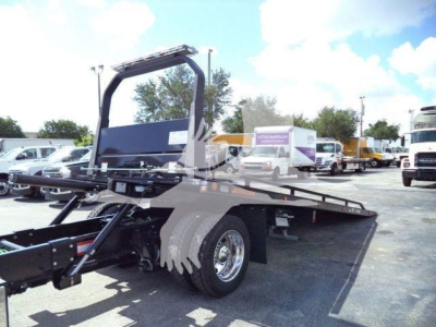 Truck rollback bed with large steel frame and wheel, showing a flatbed and hoist mechanism; background includes other trucks and a clear sky.