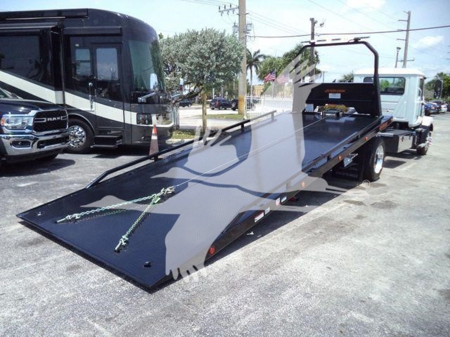 Flatbed tow truck with a hydraulic ramp in the foreground and a Mack MD6 chassis in background; heavy-duty truck with ramp and deck extended.