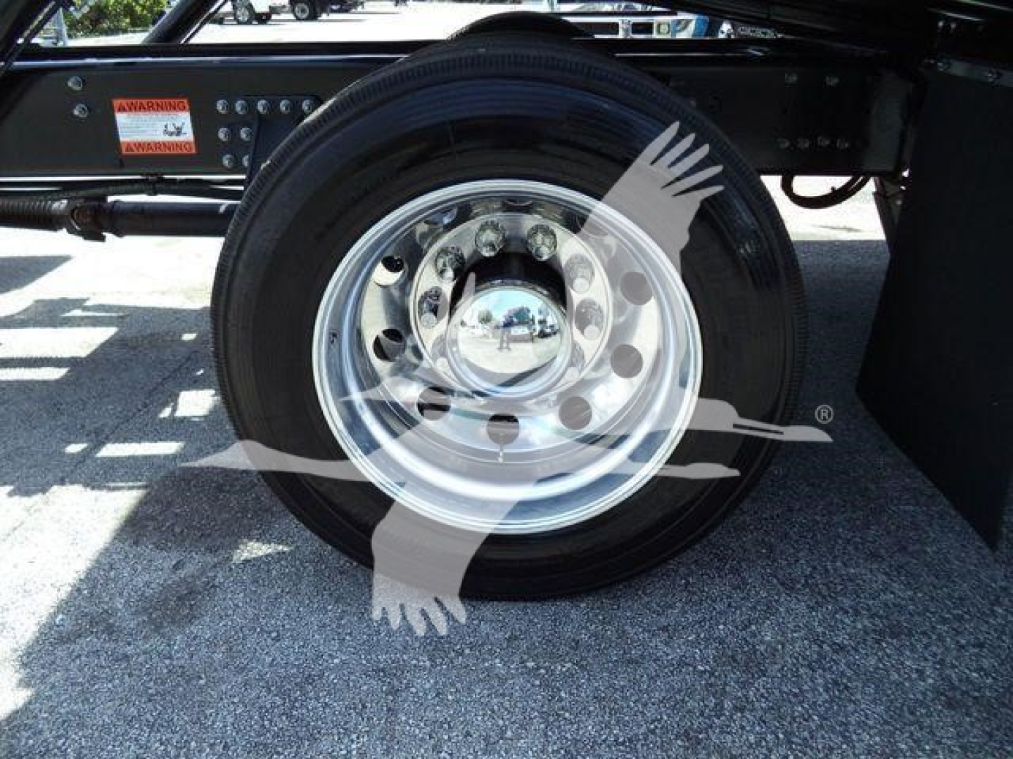 Front wheel of a 2025 Mack MD6 rollback truck showing polished steel rim and black tire with chrome hubcap; partial chassis and warning stickers visible.