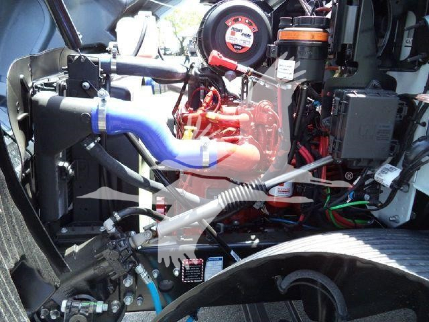 Engine bay of a 2025 Mack MD6 rollback truck showing blue coupling hoses, red engine components, wiring harnesses, and radiator area.