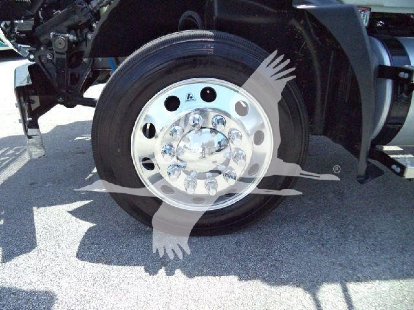 Wheel/tire assembly of a Mack MD6 truck showing polished aluminum wheel with multiple circular holes and hubcap; part of the chassis and fender visible.