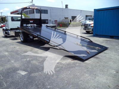 Truck with a low profile tilt-bed tow setup, hydraulic ramp/flatbed extended on a rollback tow truck with black platform and blue shipping container in background.