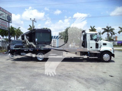 Truck-mounted rollback wrecker on a 2025 Mack MD6 chassis showing a long flatbed and a mounted crane, side view with white cab and grey bed.