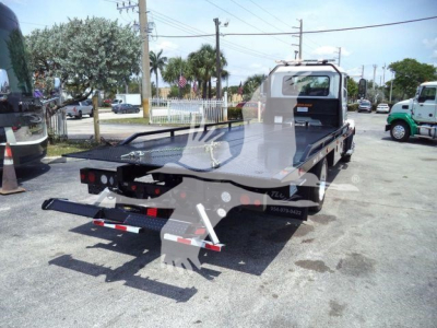Flatbed tow truck with white cab and gray flatbed, rear view showing towing yellow hooks, light bar, and platform. Yard with other trucks and palm trees visible.