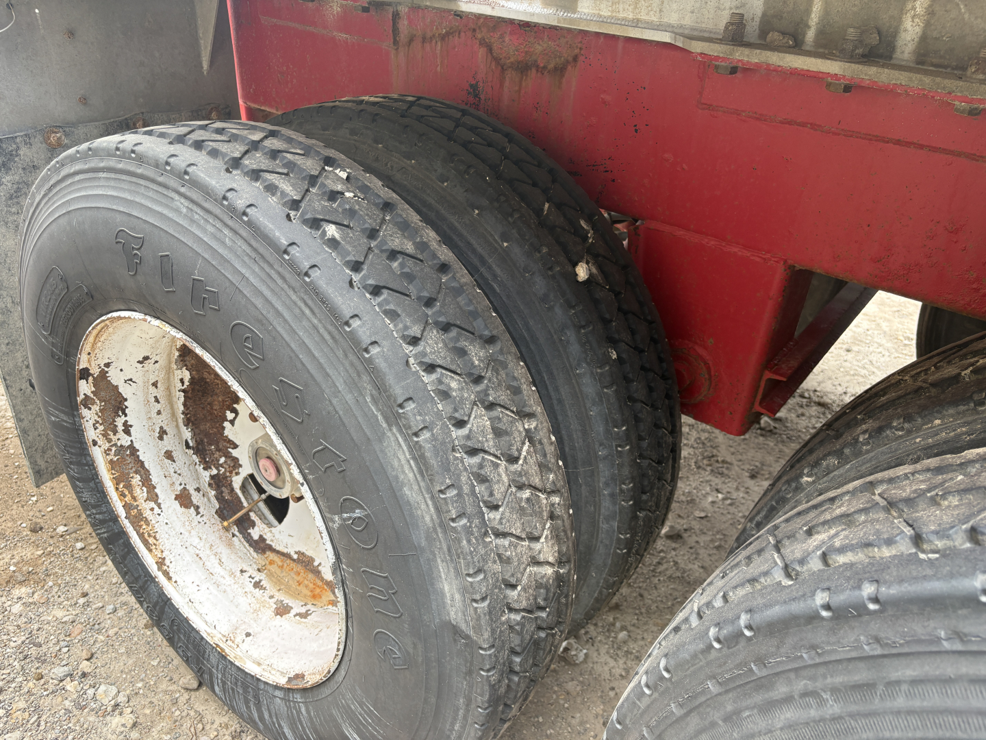 Tandem axle trailer wheel close-up with dual tires and white wheel rims, showing Vortex or brandishing text on tire, and a red trailer body in background.