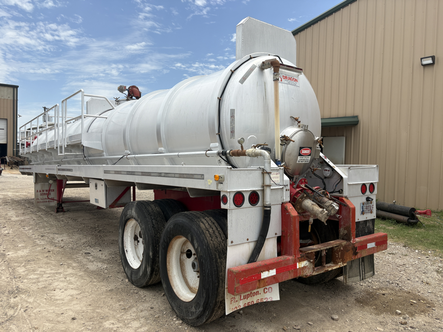 Tank trailer with white cylindrical vacuum tank attached to dual rear axles; rear valves and hoses, red hitch frame, industrial building in background. Visible text on the tank includes small labels and warning stickers.