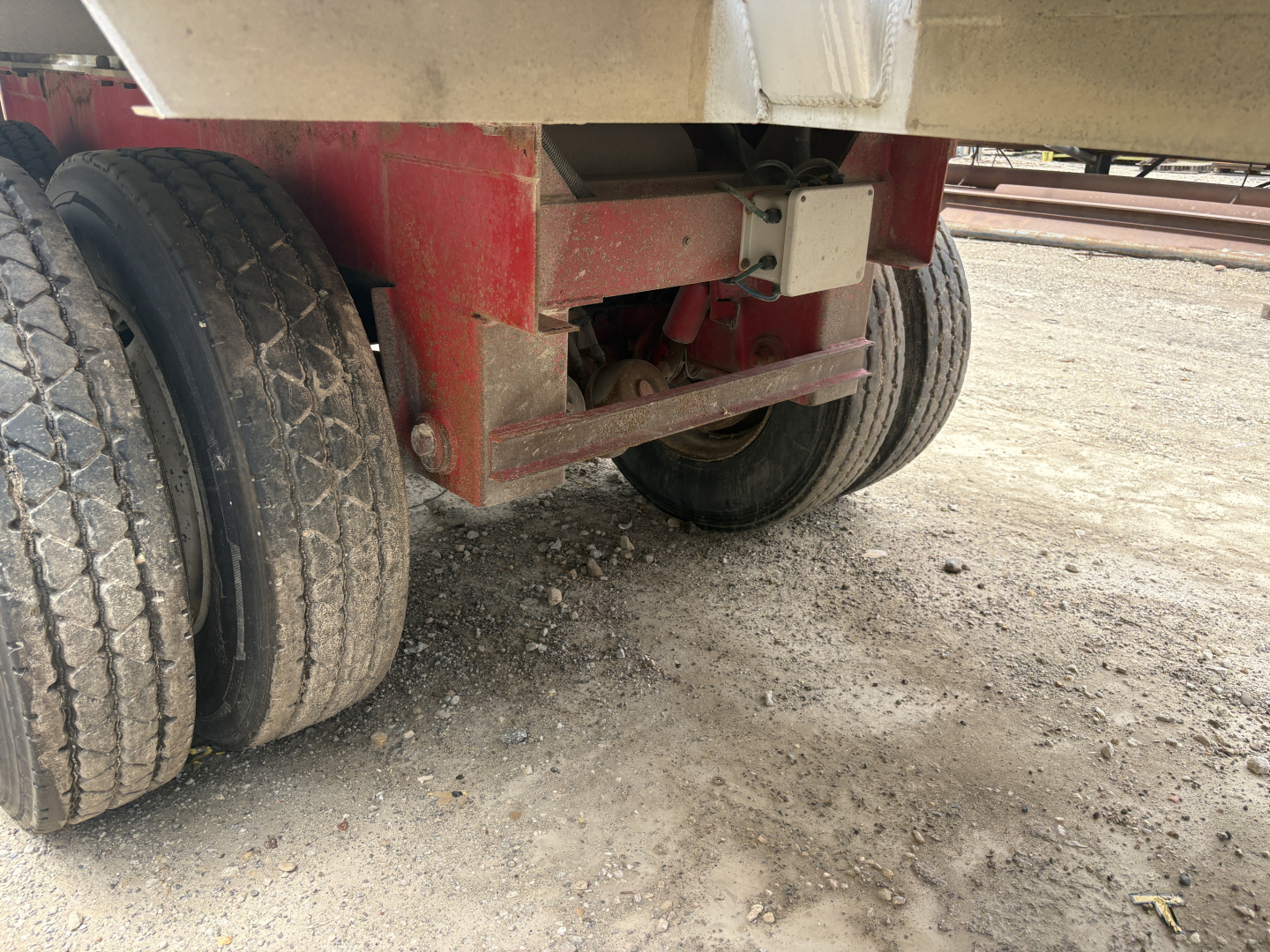 Trailer axle and undercarriage with dual rear tires, red frame and leaf spring suspension, dirt ground.