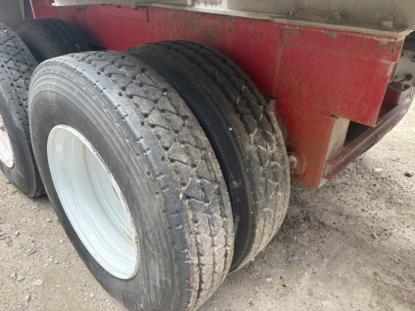 Trailer axle with two large tires mounted on white wheel rims, red trailer frame in background, gravel ground.