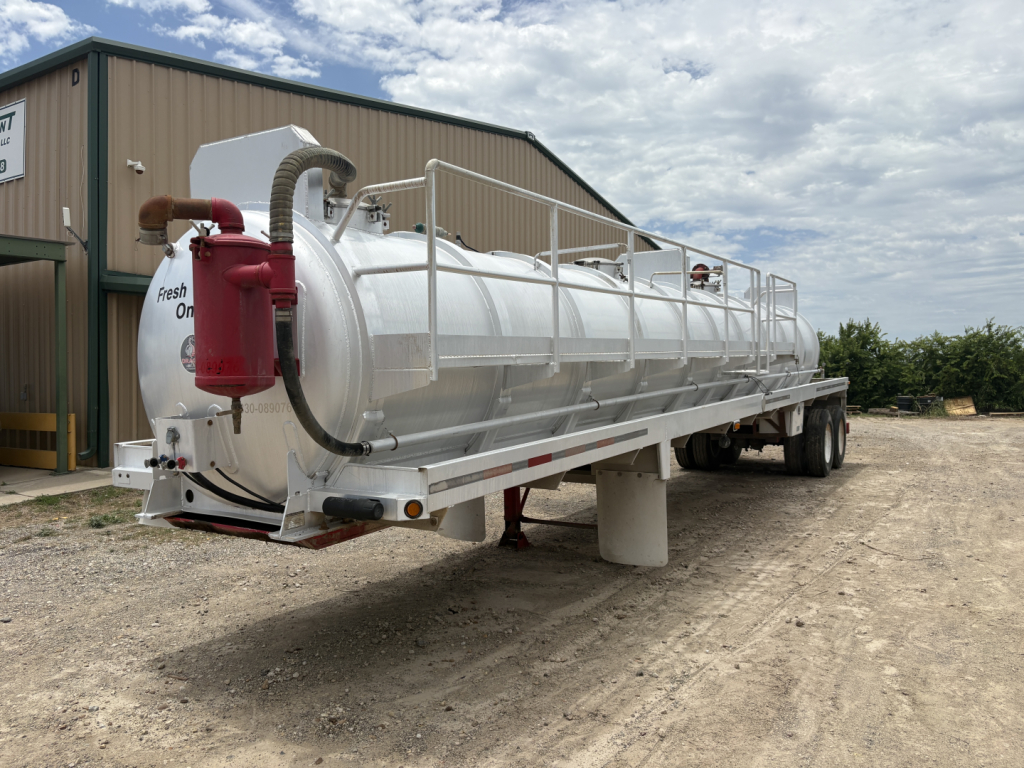 Tank trailer showing a large cylindrical stainless steel vacuum tank on a dual-axle trailer with a red control box and hoses at the rear, outdoor yard setting.