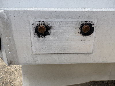 A metal plate with two rusted bolts on a trailer. The plate shows faded text: 'DBE*GON' or similar and some small numbers. The surface is metallic with dirt and wear.