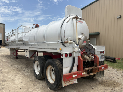 Tank trailer with white cylindrical vacuum tank attached to dual rear axles; rear valves and hoses, red hitch frame, industrial building in background. Visible text on the tank includes small labels and warning stickers.