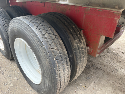 Trailer axle with two large tires mounted on white wheel rims, red trailer frame in background, gravel ground.