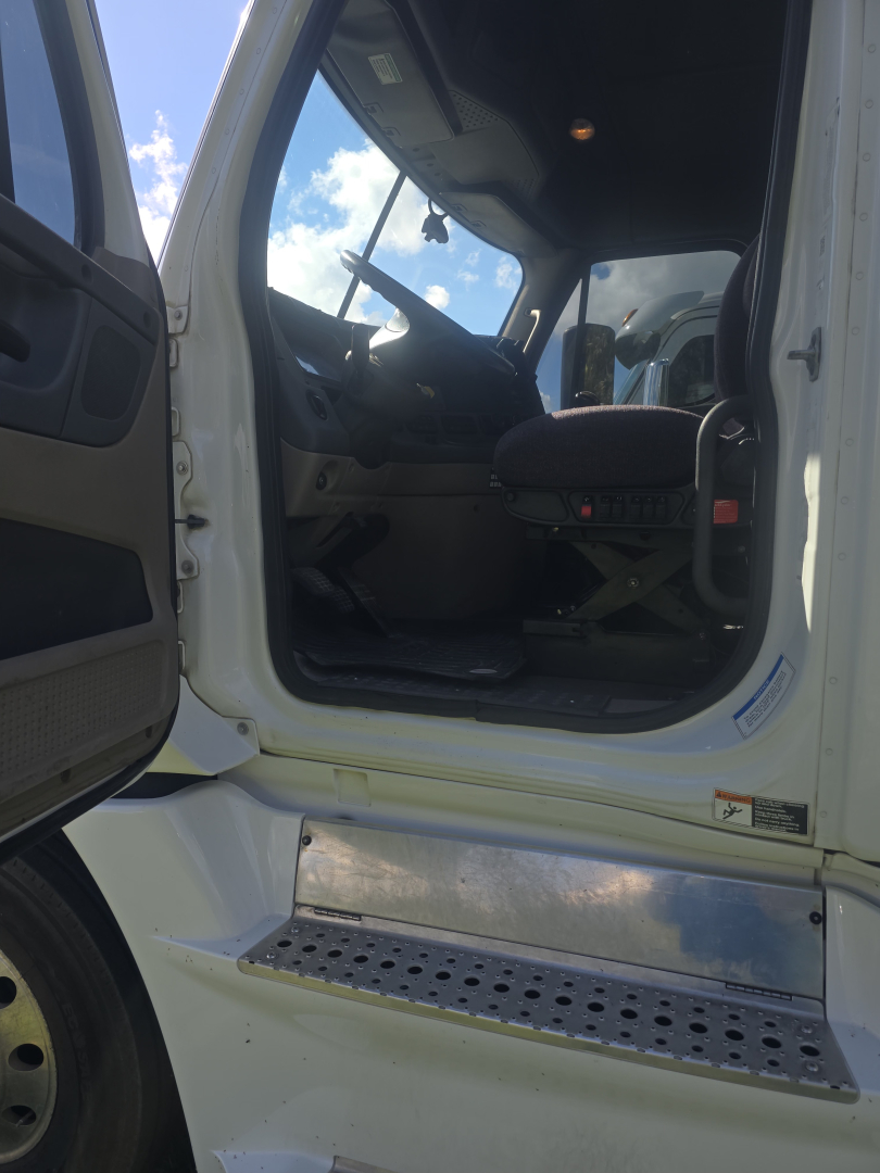 Truck cab interior visible through open door, steering wheel, driver seat, floor pedals; chrome step with perforated surface below; exterior white paint.
