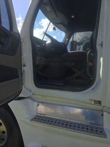 Truck cab interior visible through open door, steering wheel, driver seat, floor pedals; chrome step with perforated surface below; exterior white paint.