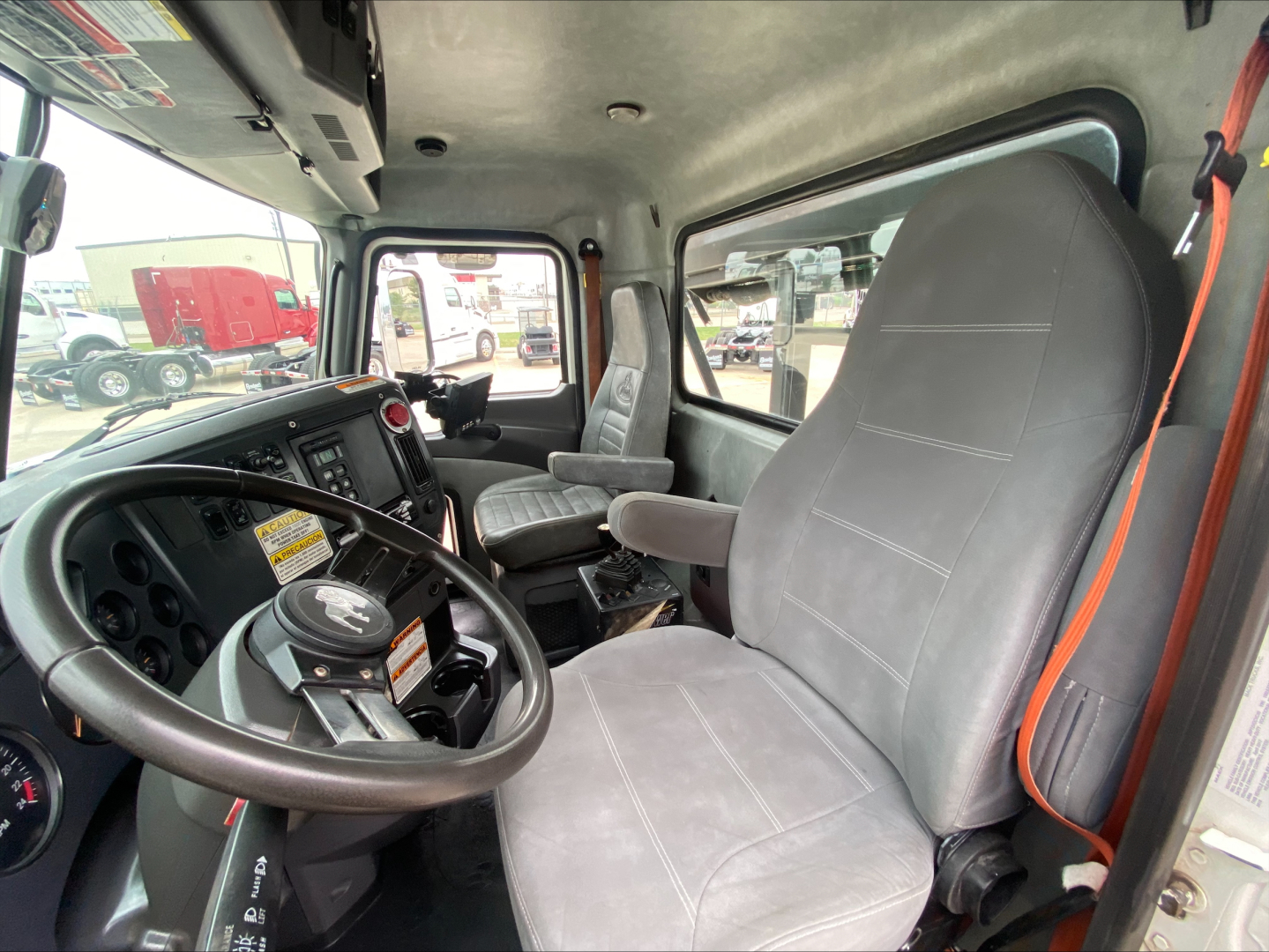 Cab interior of 2018 Mack GU533 roll-off truck showing steering wheel and dashboard, gray cloth seats, and left-side driver seat in view.