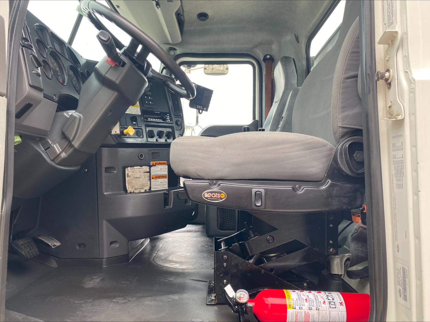 Truck cab interior of a 2018 Mack GU533 roll-off with steering wheel, dashboard, driver seat, and a red fire extinguisher mounted beneath the seat.