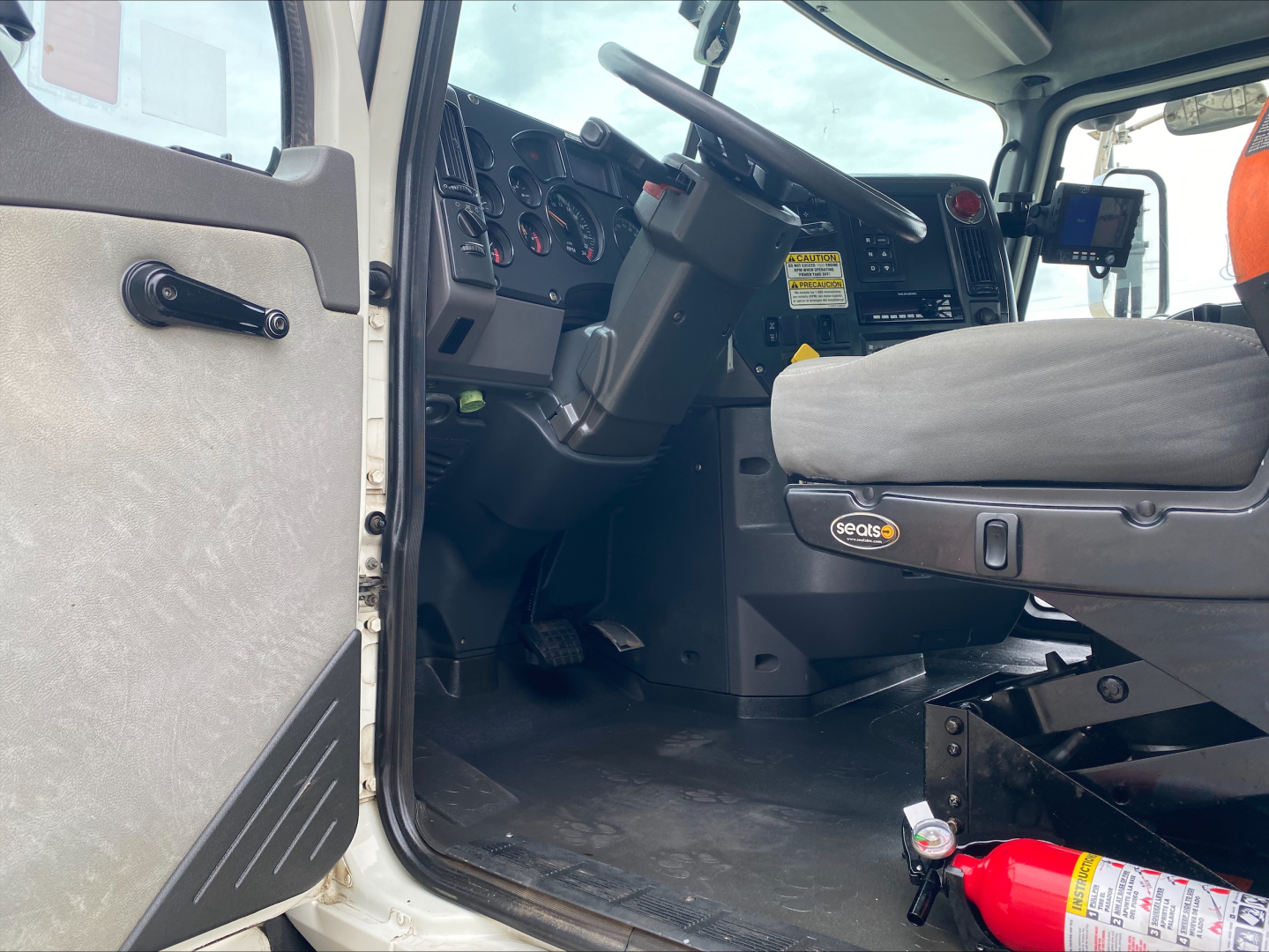Cab interior of a 2018 Mack GU533 roll-off truck showing steering column, dashboard with gauges, driver seat, and open door. Visible fire extinguisher on the floor near seat.