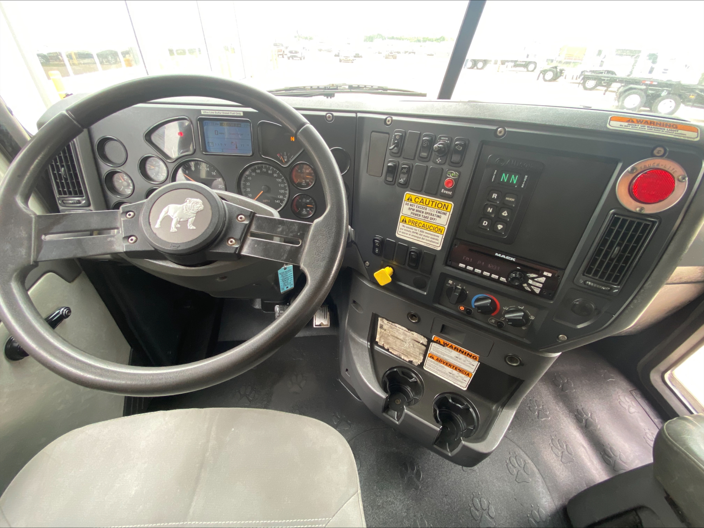 Cab interior of a 2018 Mack GU533 roll-off truck showing steering wheel, dashboard gauges, controls, and a yellow caution label.