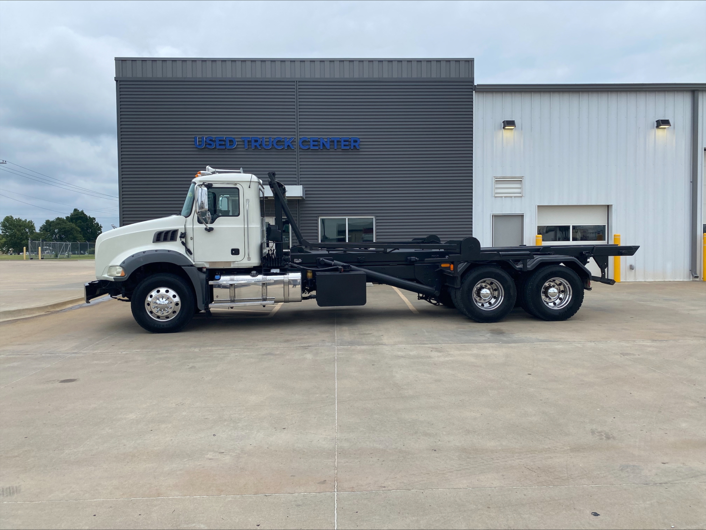 Mack GU533 truck in driveway showing side view with rolloff bed and extended chassis; factory white cab with chrome mirrors; text on building behind reads USED TRUCK CENTER.