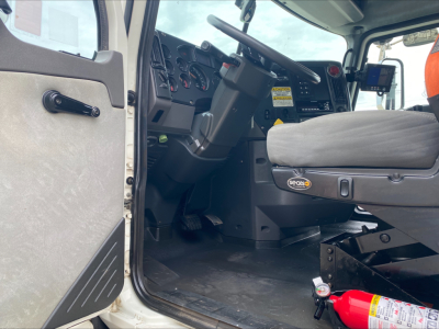 Cab interior of a 2018 Mack GU533 roll-off truck showing steering column, dashboard with gauges, driver seat, and open door. Visible fire extinguisher on the floor near seat.