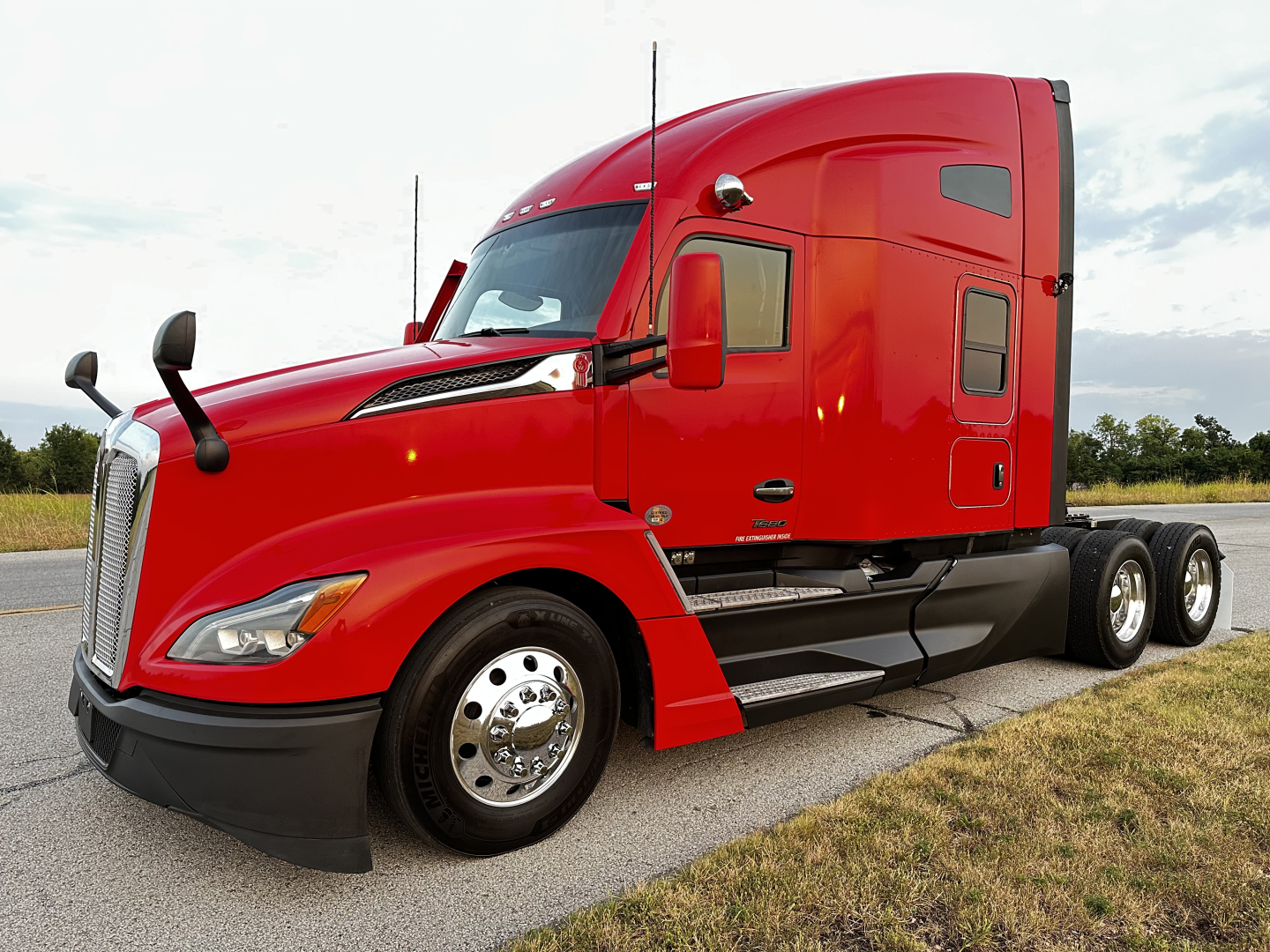 Kenworth T680 conventional sleeper tractor in red, side view showing truck cab with door, mirrors, fuel tanks, and dual rear axles.
