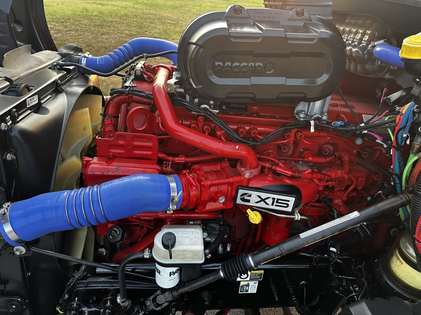 Kenworth T680 engine bay with red 6-CTA engine, blue hoses, and visible air intake cover showing Vol. 2X15 badge; various wiring and reservoirs present.