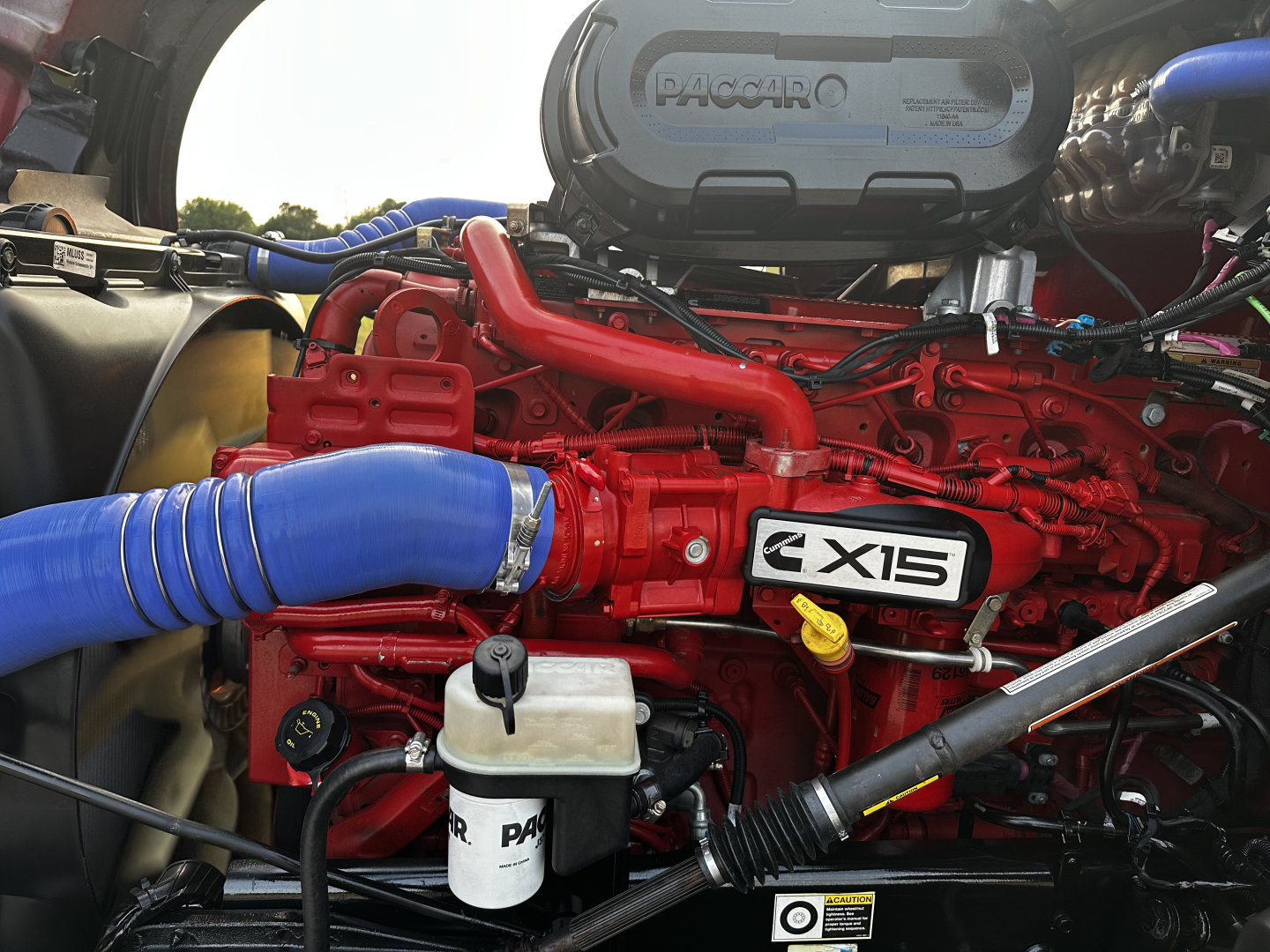 Kenworth T680 engine bay showing red X15 engine with blue intercooler hose and Paccar badge visible.