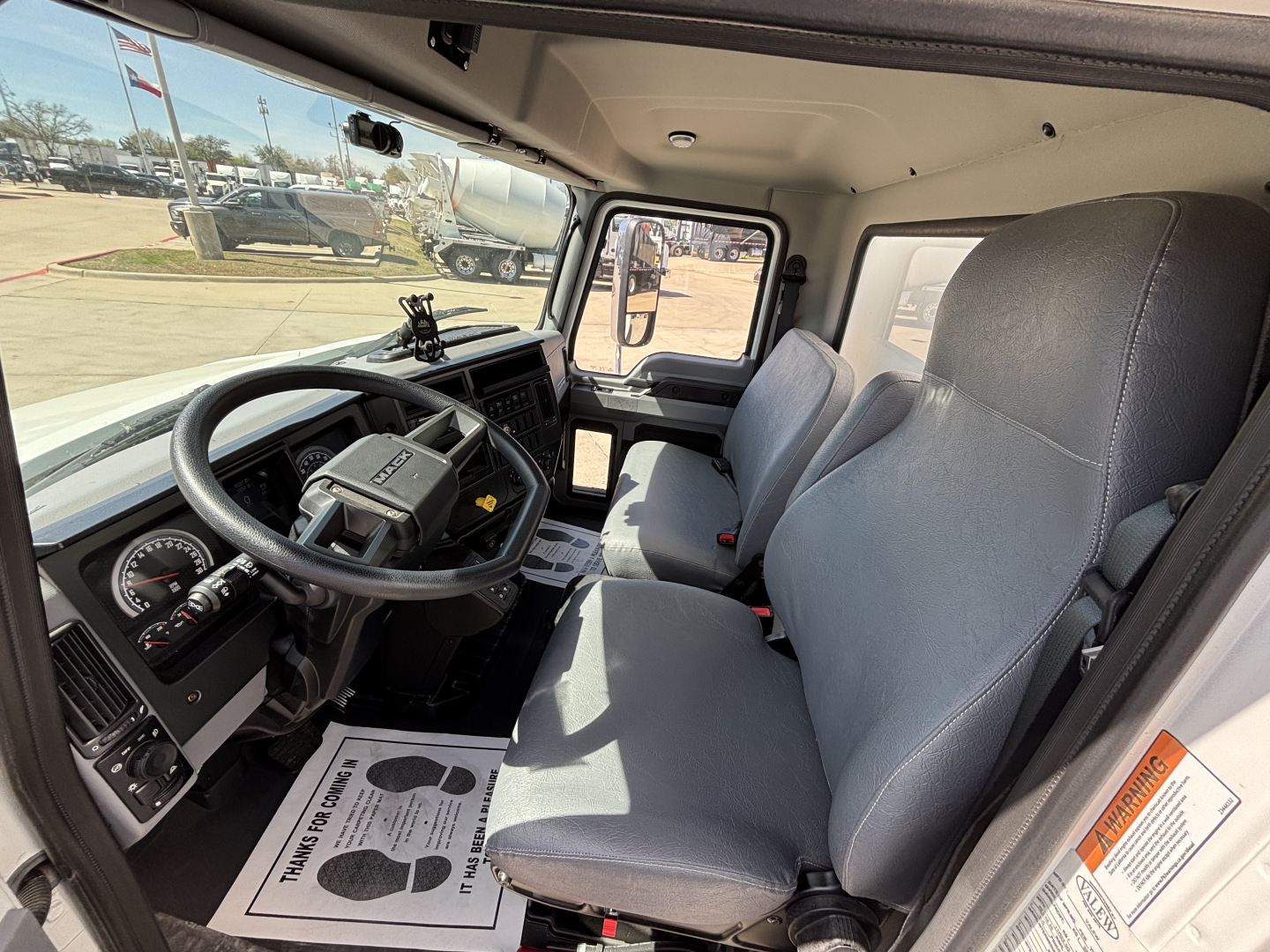 Cab interior of a 2025 Mack MD7 truck showing steering wheel, dashboard, and two gray seats in daylight.